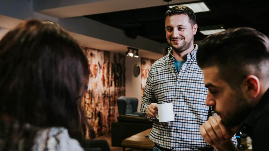 A man in a plaid shirt holding a coffee mug and chatting with coworkers.