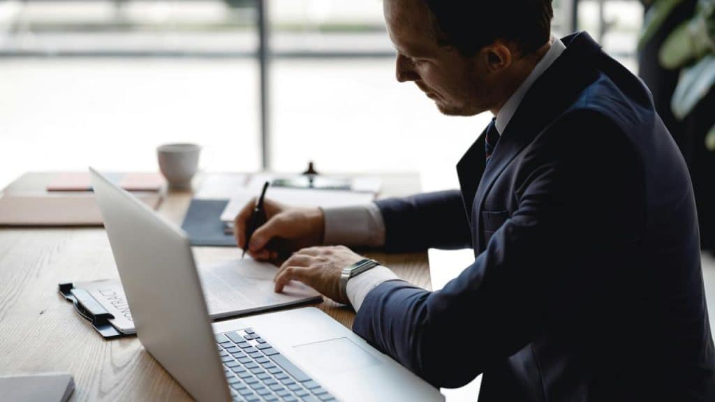 A man in a suit signing a document at a desk beside a laptop.