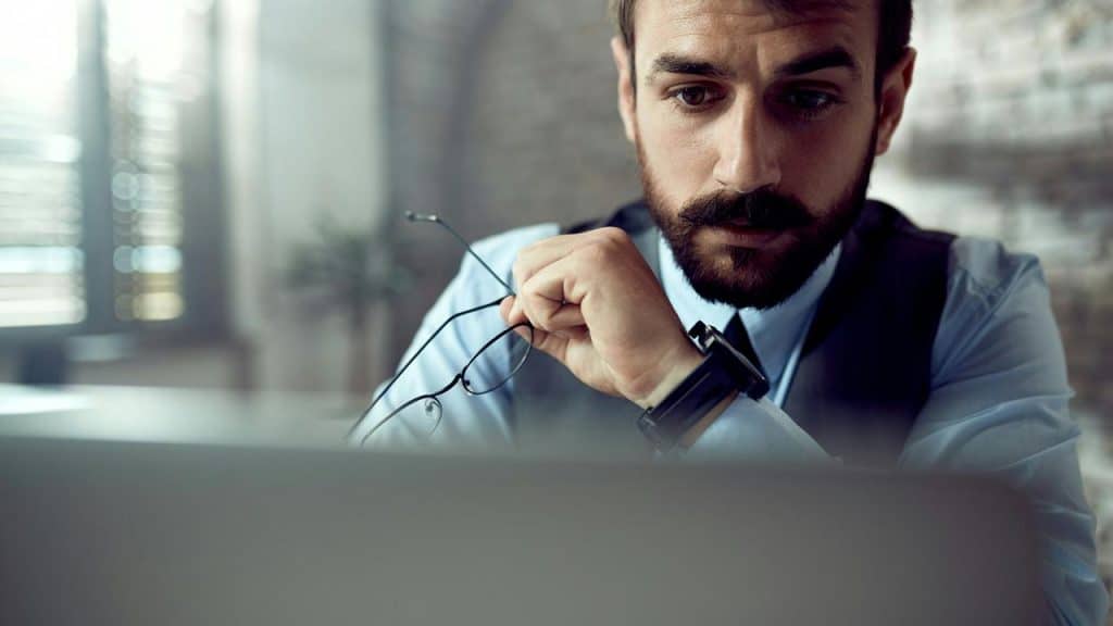 A bearded man holding his glasses while intently viewing a laptop.
