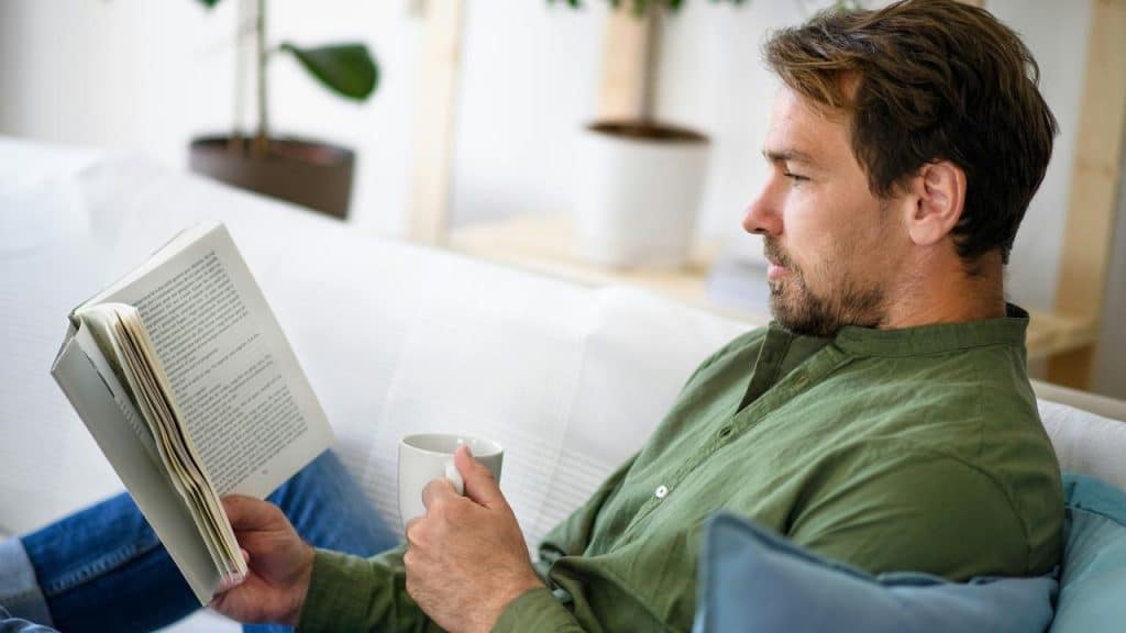 A man reading a book on a couch while holding a mug.