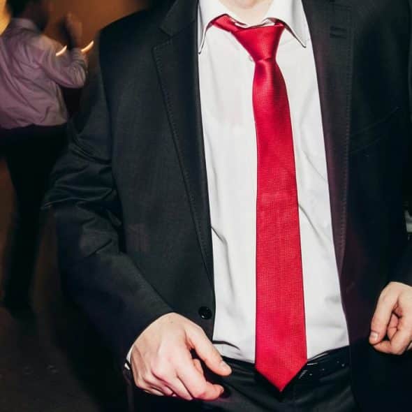 A man in a dark suit and bright red tie standing indoors at a formal event.