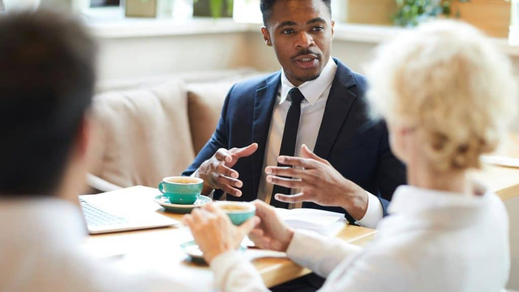 A man in a dark suit gesturing while talking over coffee with two colleagues in a café.