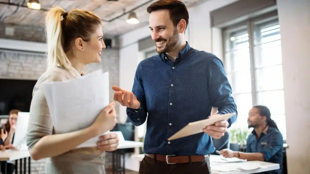 A man with a tablet talking to a woman holding papers in an office.