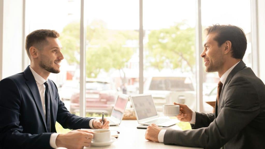 A man in a dark suit and a man in a gray suit talking over coffee with laptops on a table.