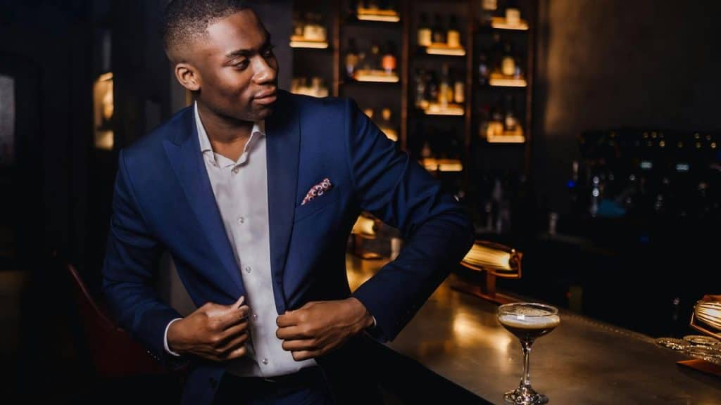 A well-dressed man in a navy suit adjusting his jacket at a dimly lit bar counter with a cocktail nearby.