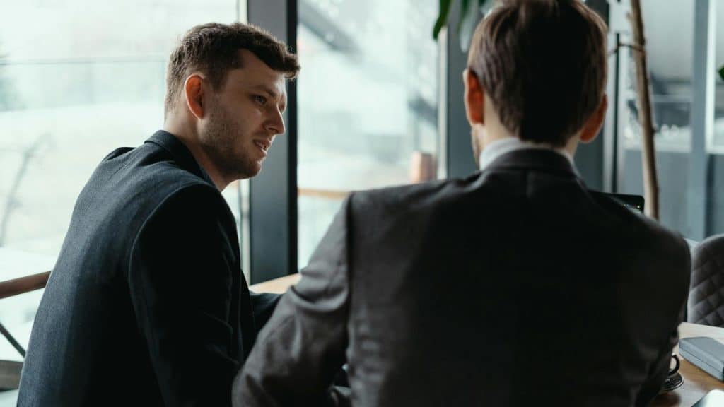 A man in a dark suit speaking to a colleague by a window.