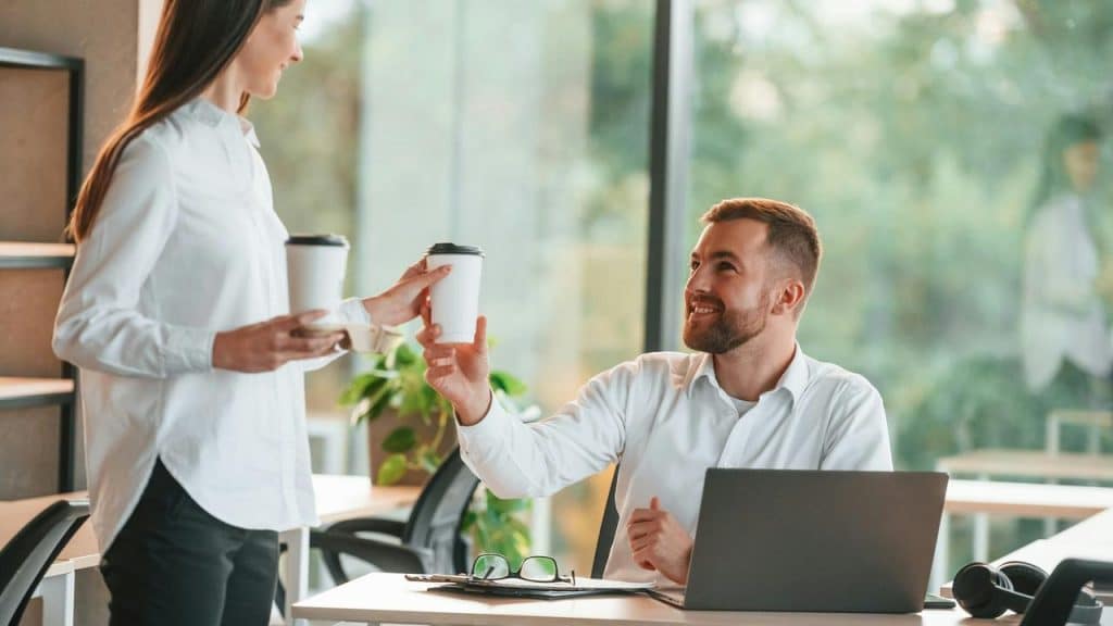 A woman handing a coffee cup to a seated man at his desk.