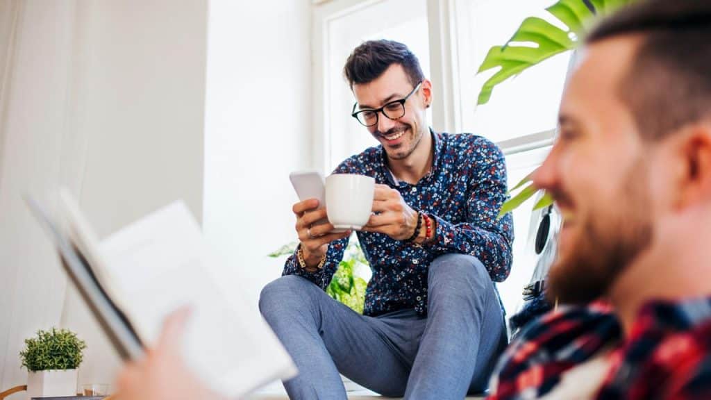 A man with glasses holding a coffee mug and checking his phone while sitting casually.