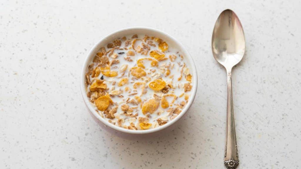 A bowl of cornflakes and granola in milk on a speckled countertop, with a silver spoon beside it.