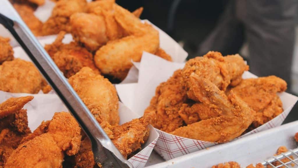 A close-up of golden, crispy fried chicken pieces nestled in paper trays with metal tongs.