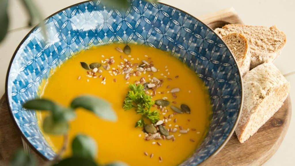 A bowl of orange soup topped with seeds and parsley beside sliced bread.