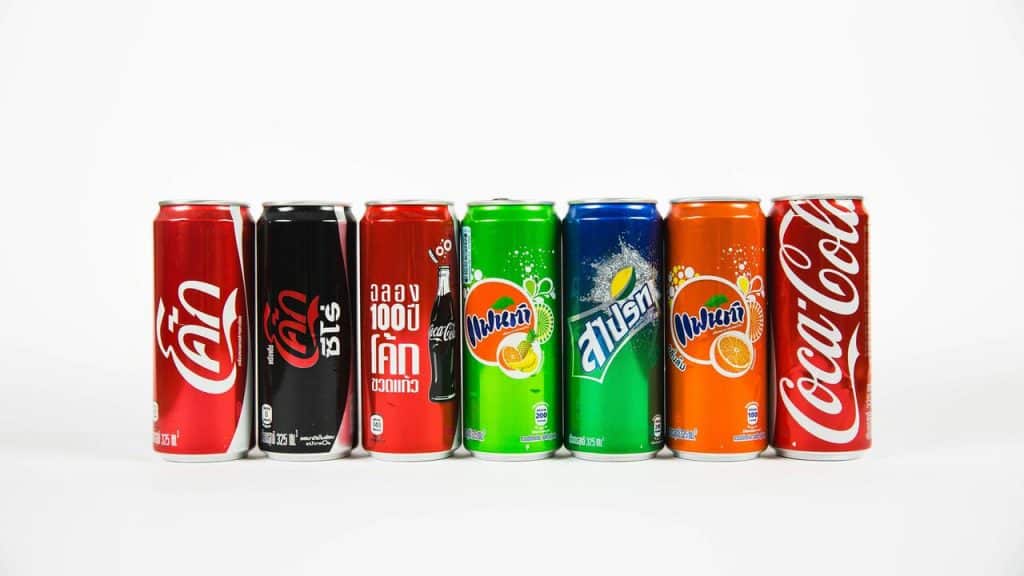 A lineup of assorted soda cans, including Coca-Cola, Sprite, and Fanta variants, against a white background.