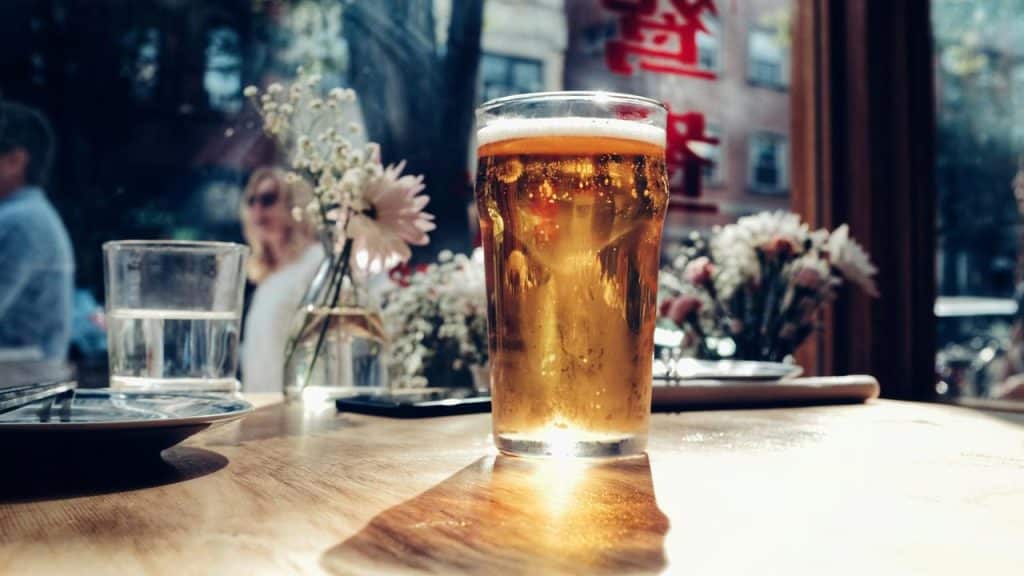 A pint of golden beer on a sunlit wooden table with blurred flowers in the background.