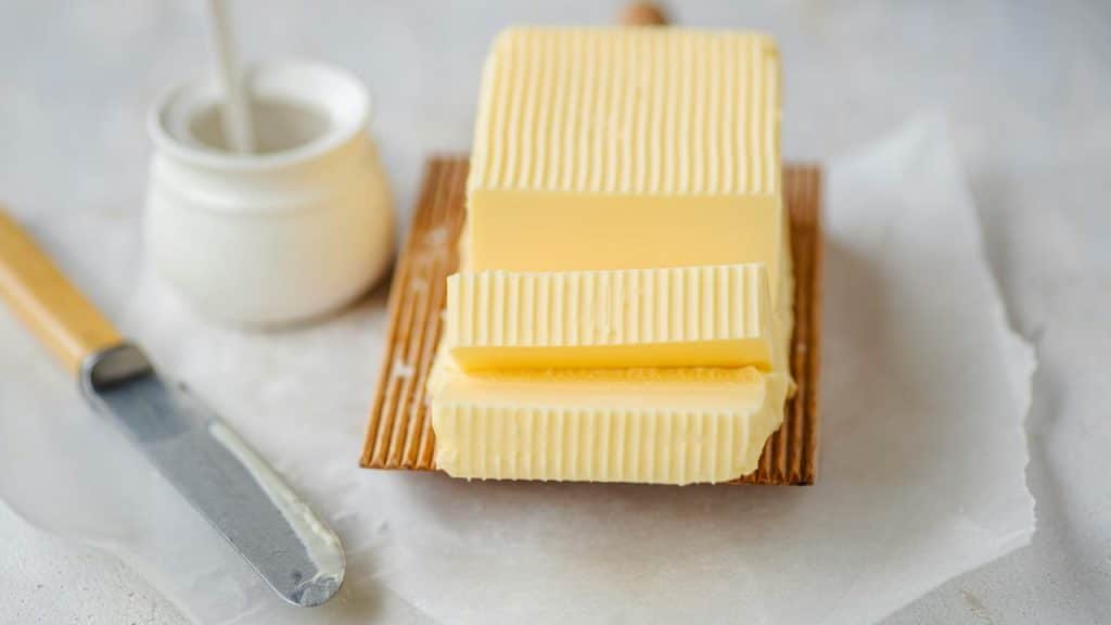 A block of butter sliced into ridged slabs on parchment paper with a knife nearby.