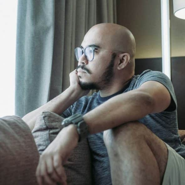 A bald man with a beard and glasses sits on a couch, looking pensively out a window.