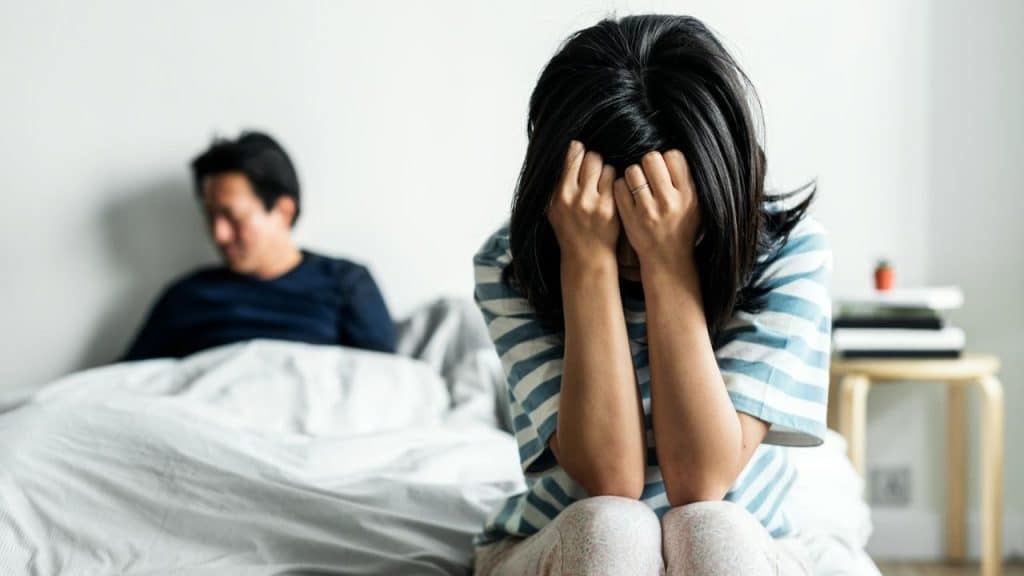A woman sits on a bed, covering her face with her hands, while a man sits in the background.