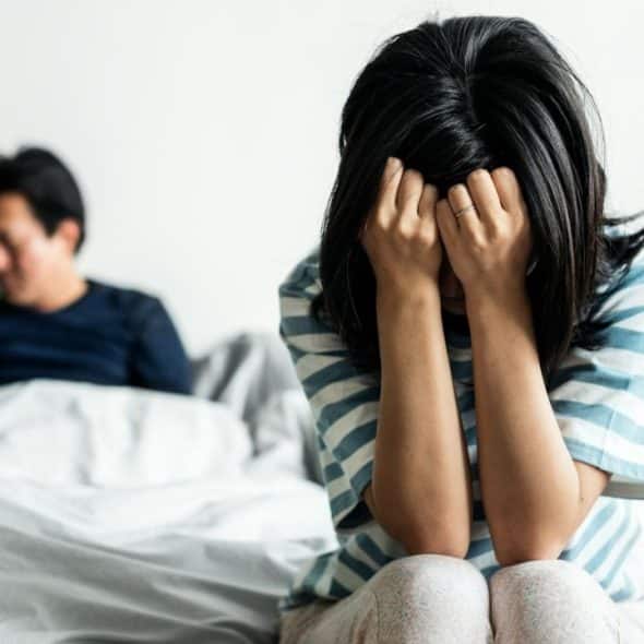 A woman sits on a bed, covering her face with her hands, while a man sits in the background.