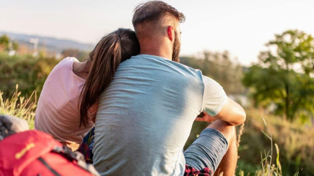 A man and a woman sit outdoors, looking away, with the woman leaning on the man's shoulder.