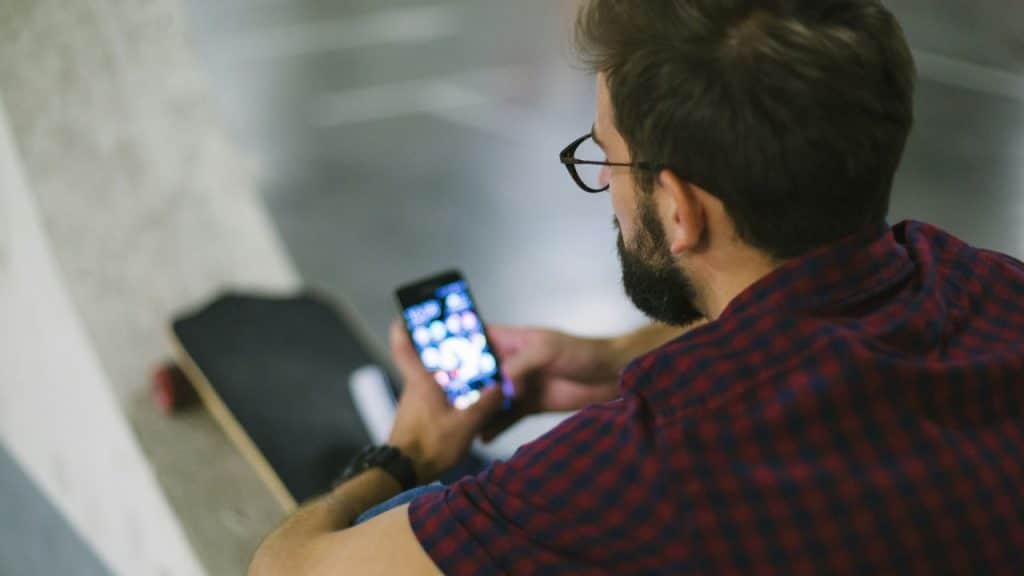 A bearded man wearing glasses and a plaid shirt looks at his phone, with a skateboard nearby.