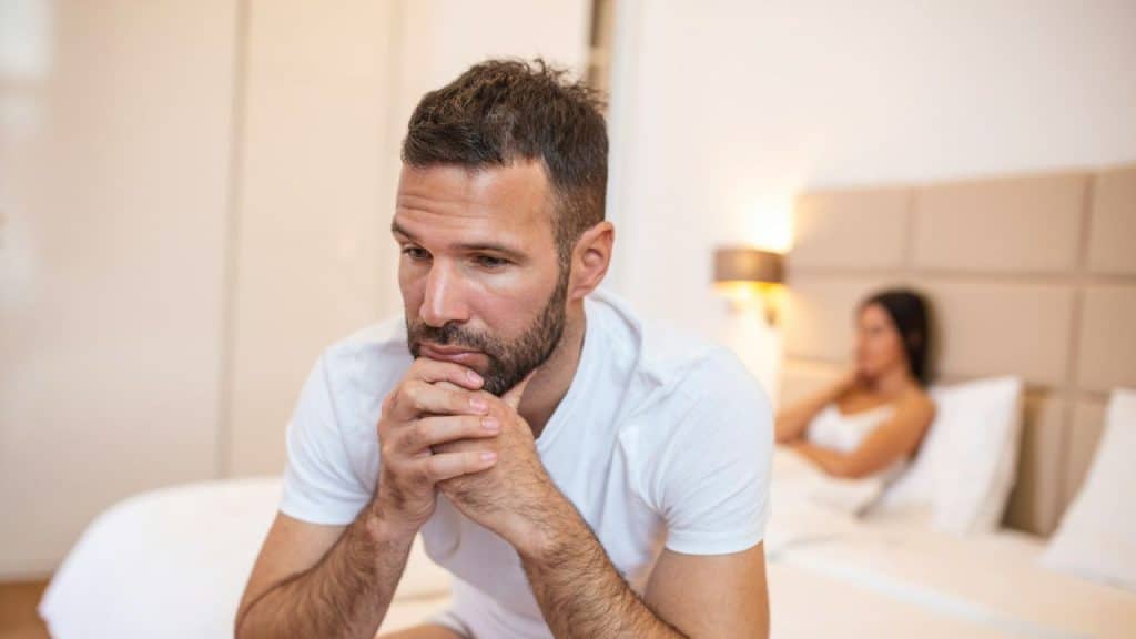 A thoughtful bearded man in a white shirt sits on a bed, with a woman in the background.