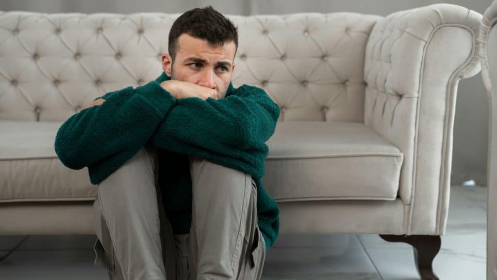 A man in a green fleece and light pants sits on the floor, hugging his knees, next to a couch.