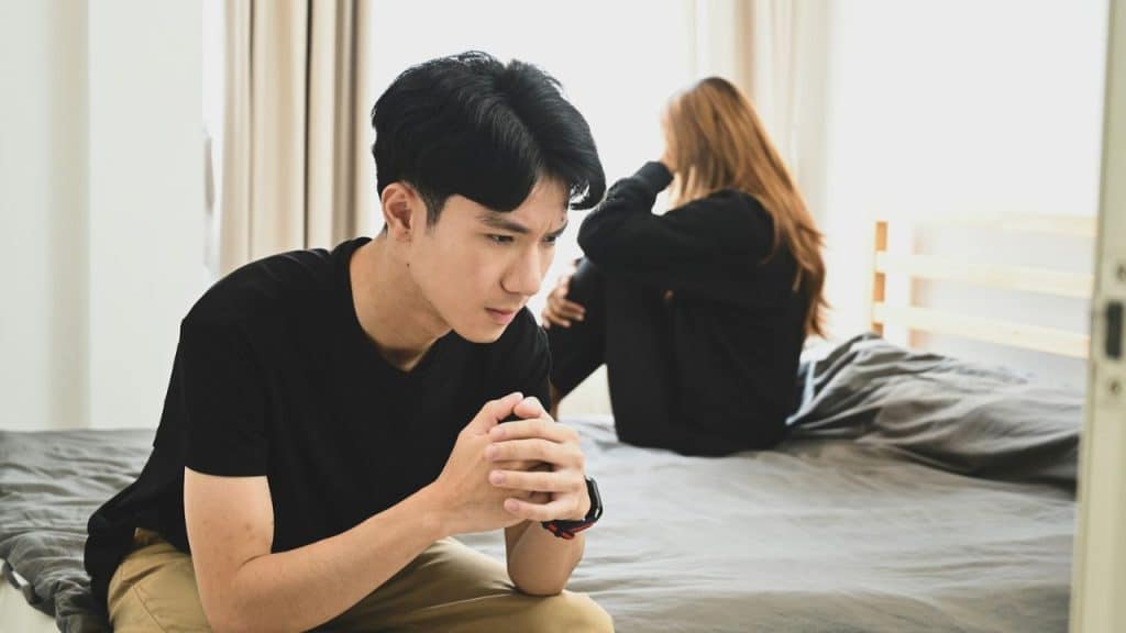 A thoughtful man sits on a bed, looking down, while a woman sits with her back to him in the background.