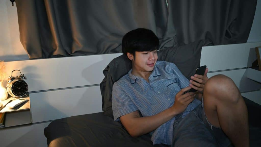A smiling man sits up in bed, looking at his phone, with an alarm clock on a nightstand.