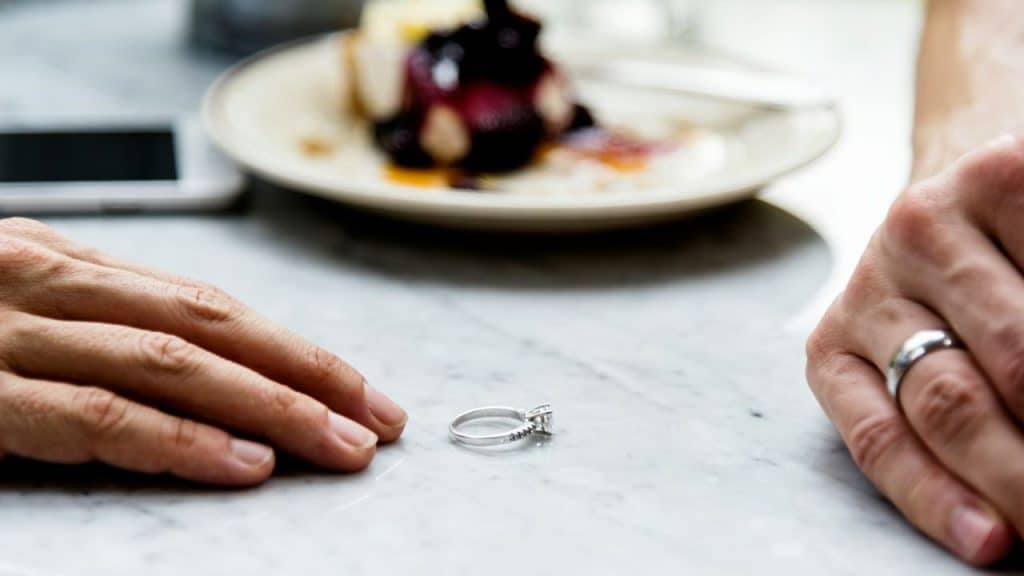 A diamond ring lies on a marble table between two hands, one wearing a band.