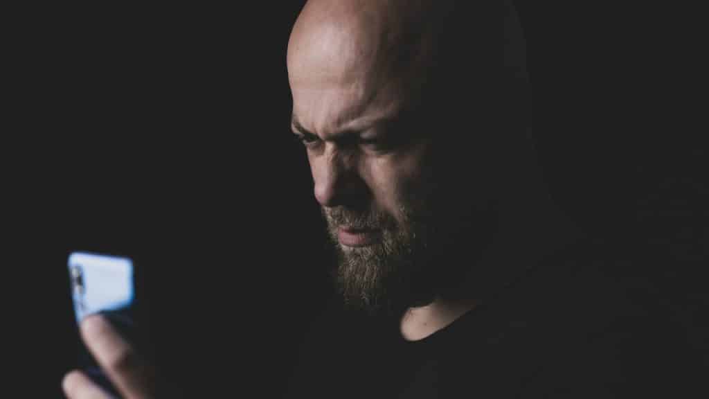 A bald, bearded man with a stern expression looks at a phone in a dark room.