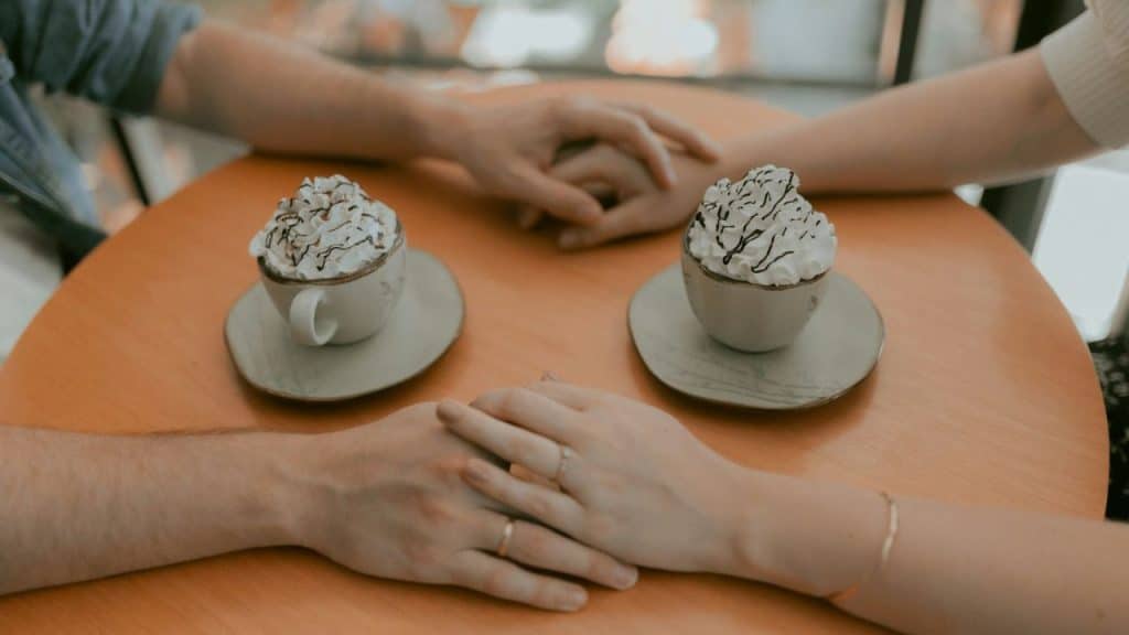 A pair of hands with rings holding, and another pair of hands separate, on a table with two whipped cream drinks.