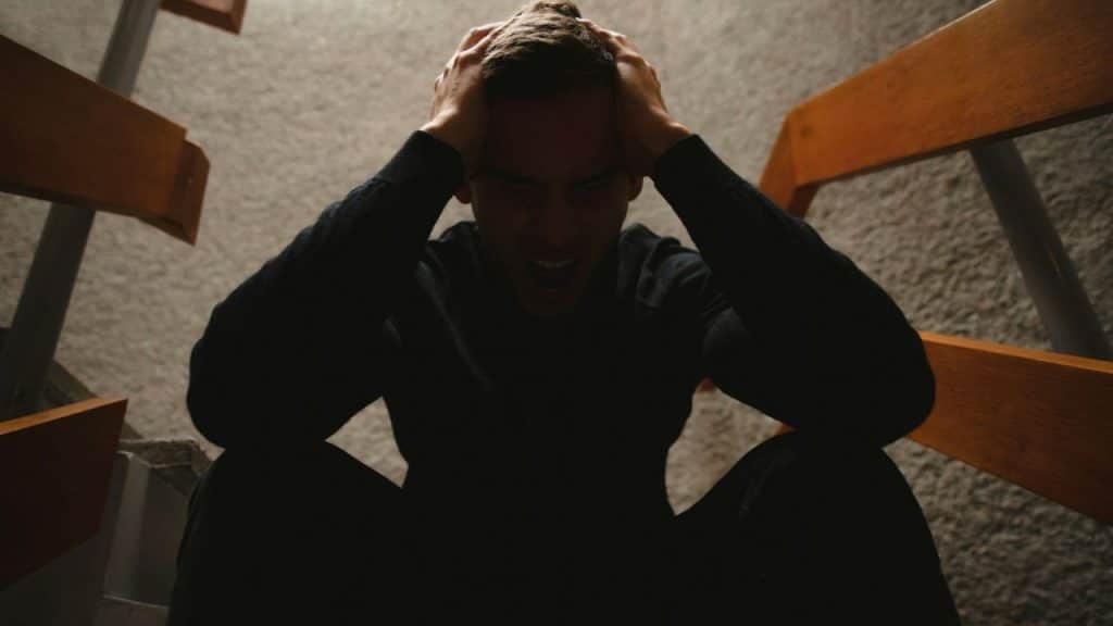A distressed man is seated, clutching his head with both hands, in a dimly lit setting.