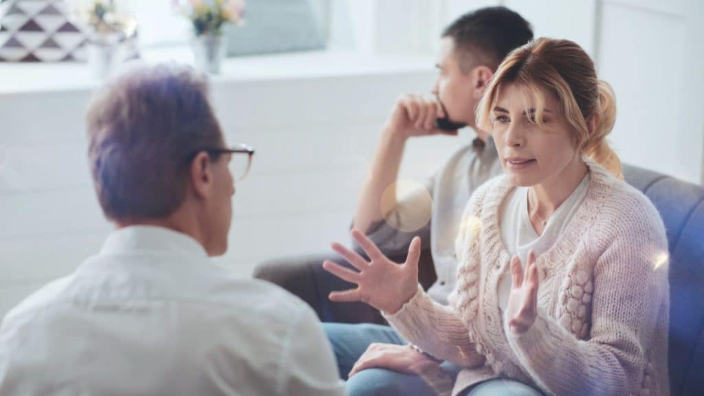 A woman gestures while talking to a man with his back to the camera, and another man sits behind her.