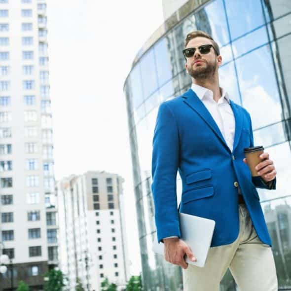 A man in a blue blazer and sunglasses walks confidently with a laptop and coffee cup.