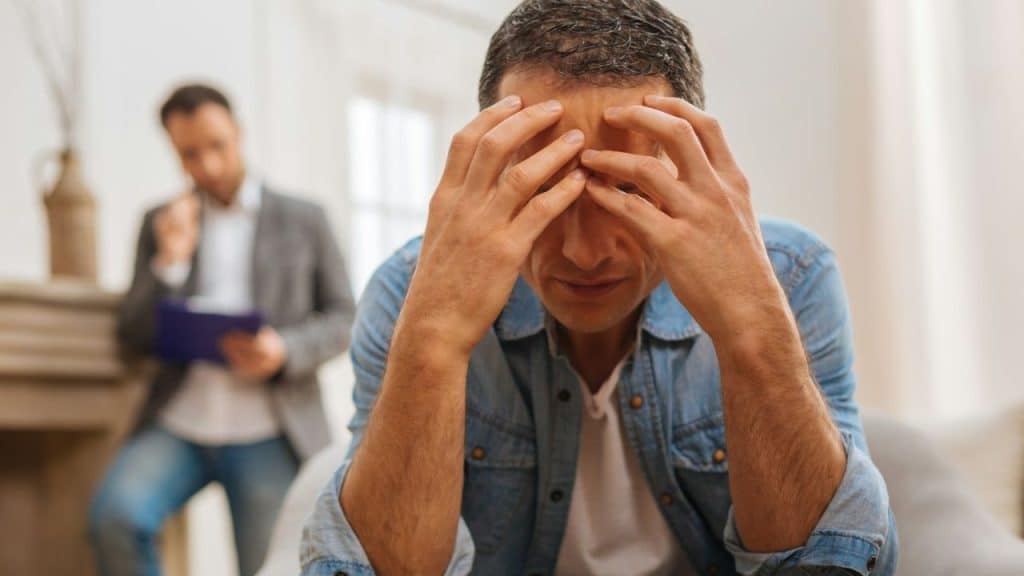 A man in a denim shirt holds his head in his hands, looking distressed, while another man sits in the background.