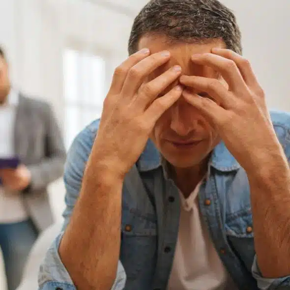 A man in a denim shirt holds his head in his hands, looking distressed, while another man sits in the background.