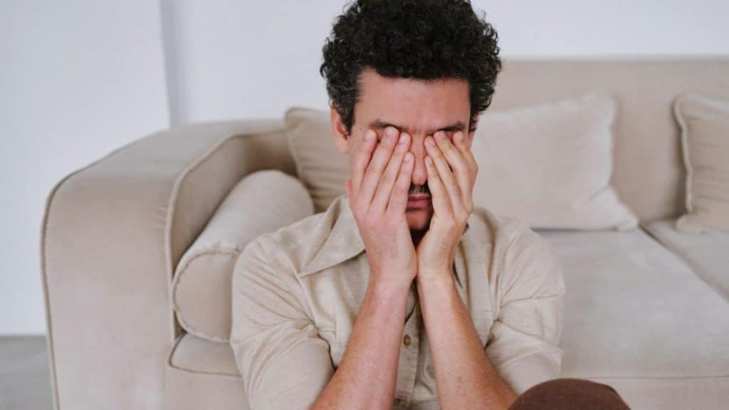 A man with curly hair sits on the floor, covering his face with his hands.