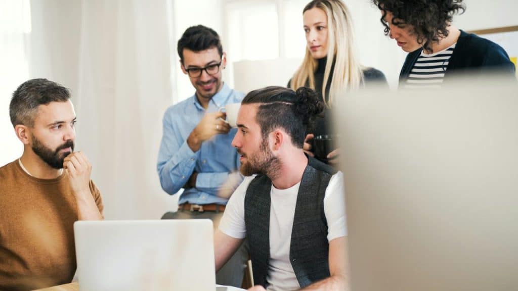 A group of five diverse individuals are gathered around a laptop in an office setting.