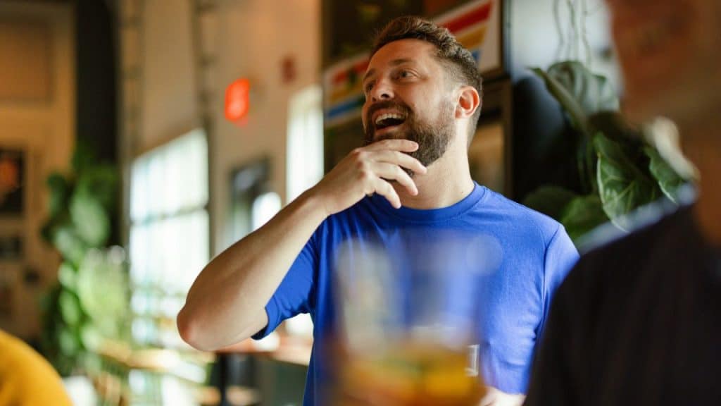 A smiling man with a beard, wearing a blue shirt, stands indoors.