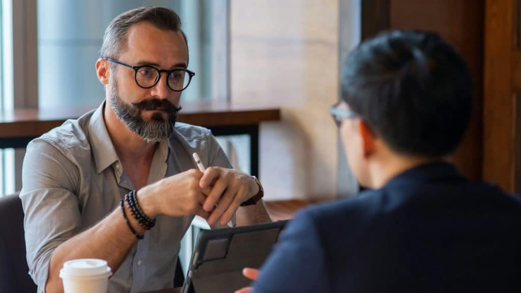 A man with a beard, mustache, and glasses talks to another person over a tablet and coffee.