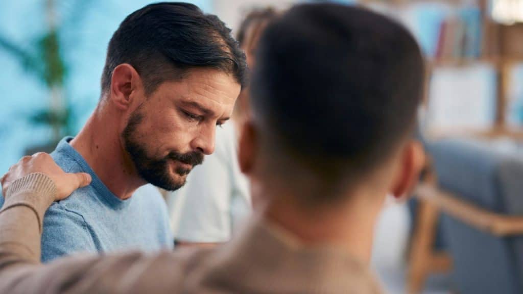 A man with a beard looks down, while another person's hand rests on his shoulder.