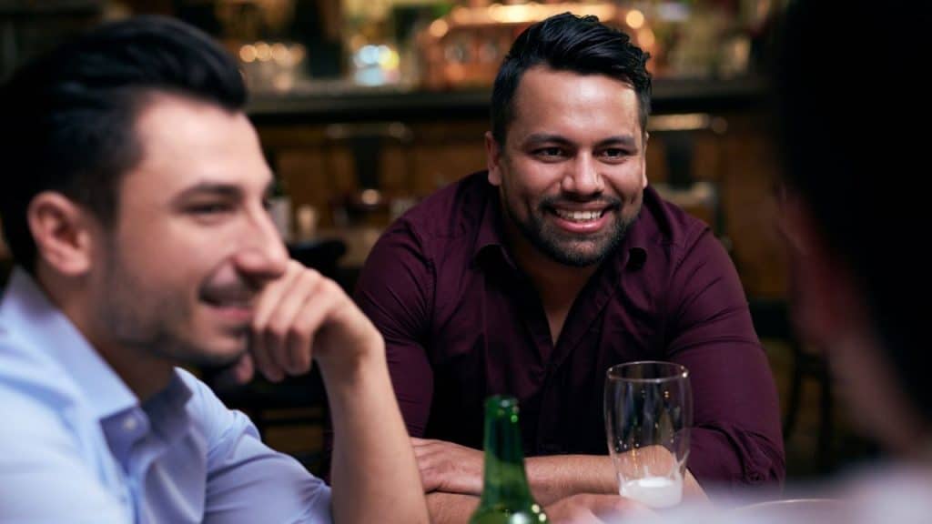 A smiling man with a beard sits at a table with other men, with a beer bottle in front.