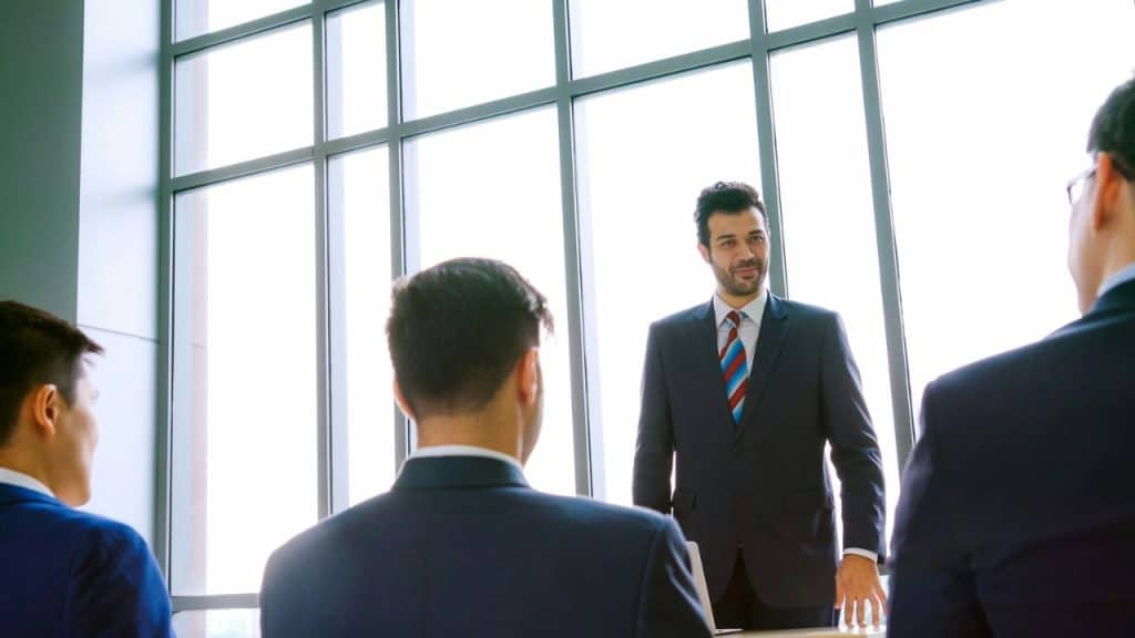A man in a suit and striped tie stands in front of a window, looking at three men with their backs to the camera.