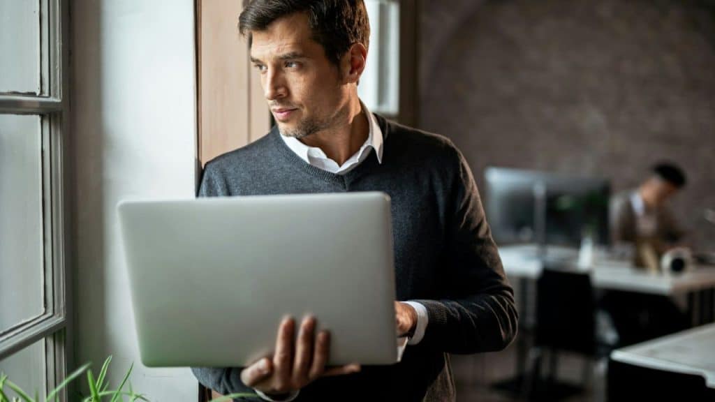 A man in a dark sweater and white collared shirt holds a laptop, looking out a window.