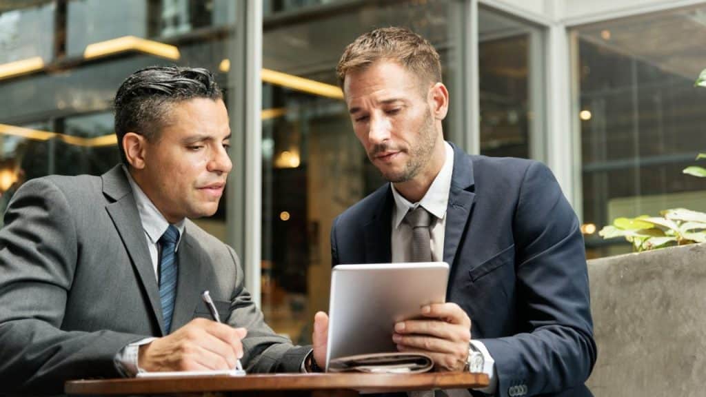 Two men in suits sit at a table; one writes on paper while the other holds a tablet.