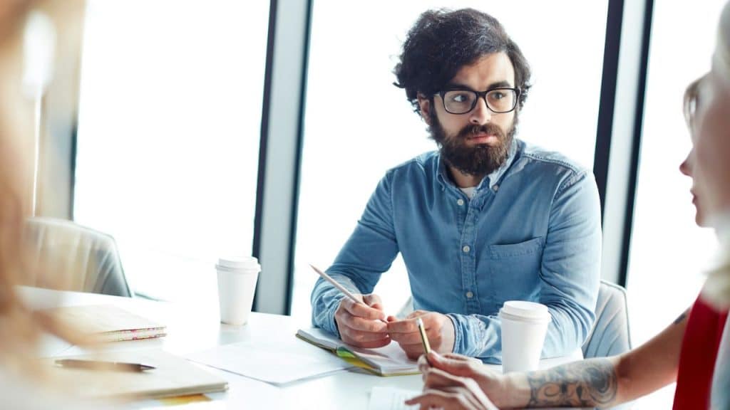 A bearded man in glasses holds a pencil, looking intently at someone off-camera.