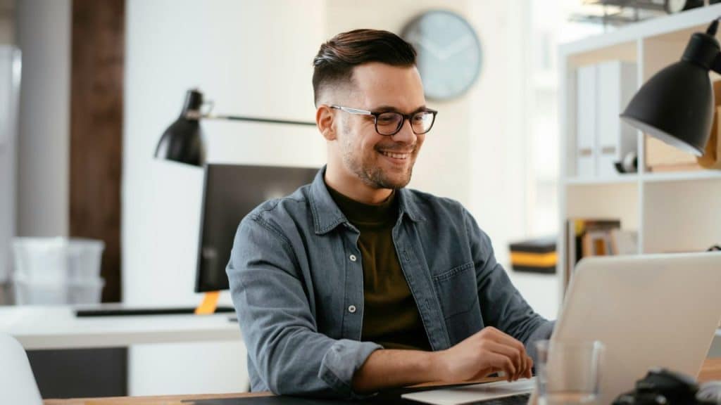 A smiling man with glasses and a beard works on a laptop at his desk.