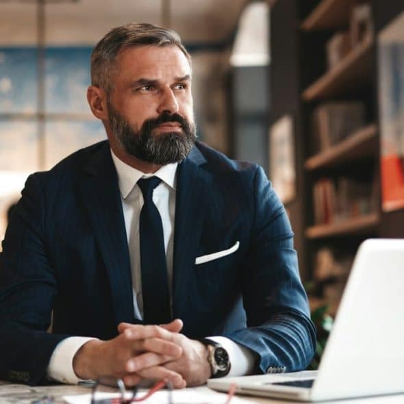 A distinguished, bearded man in a suit looks thoughtfully to the right, sitting at a table with a laptop.