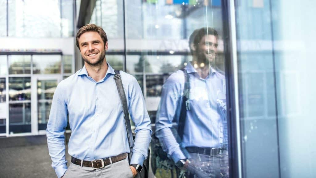 A smiling man in a blue shirt and grey pants stands with his hands in his pockets, reflecting in a window.