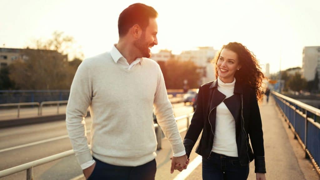 A smiling couple walks hand-in-hand on a bridge, looking at each other.