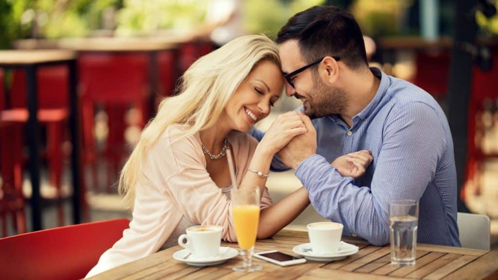 A man and a woman are seated at an outdoor cafe table, holding hands and smiling at each other.